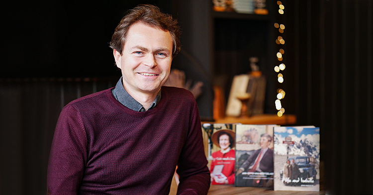 shows a white man with brown hair and a red shirt standing in front of books